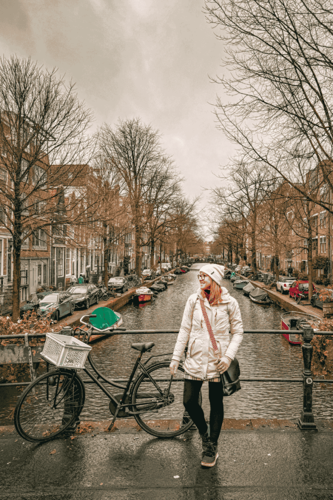 Kate smiles while standing on a bridge next to a bike in Amsterdam, with a tree-lined canal and rows of parked boats stretching behind her.