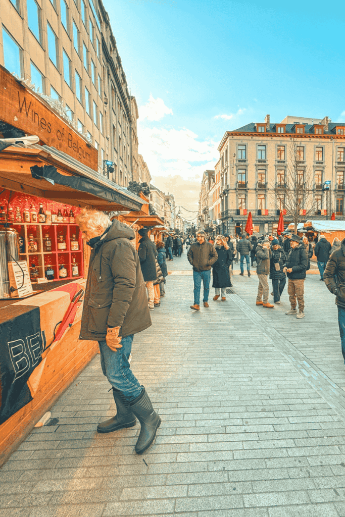 Visitors browse cozy wooden stalls at a festive Brussels street market, with a "Wines of Belgium" booth in the foreground and crowds enjoying the winter atmosphere.