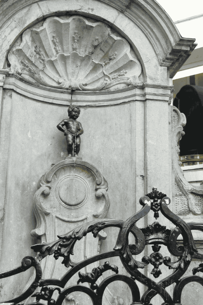 The Manneken Pis statue in Brussels stands behind a decorative wrought iron gate, nestled in its stone niche with a scalloped shell design above.