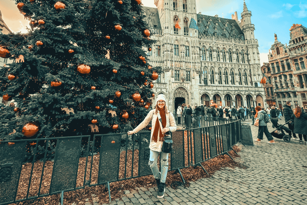 Kate stands smiling in front of a massive decorated Christmas tree at Grand Place in Brussels, with the ornate Town Hall and festive crowds in the background.