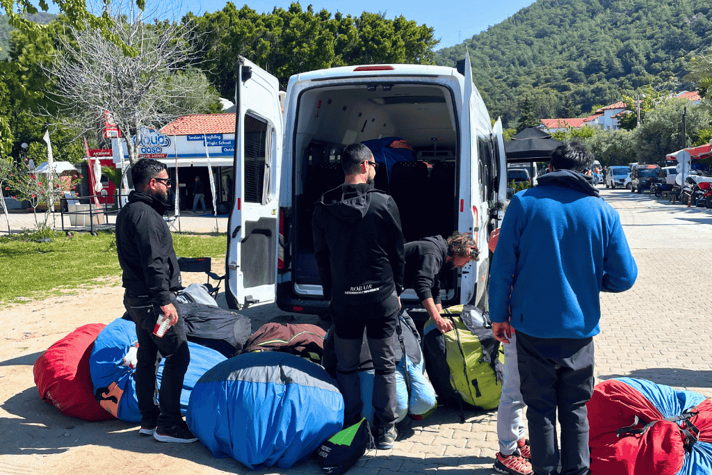 A group of paragliding crew members pack colorful parachutes into a van under bright morning sun in Ölüdeniz, Turkey, preparing for a trip up the mountain.