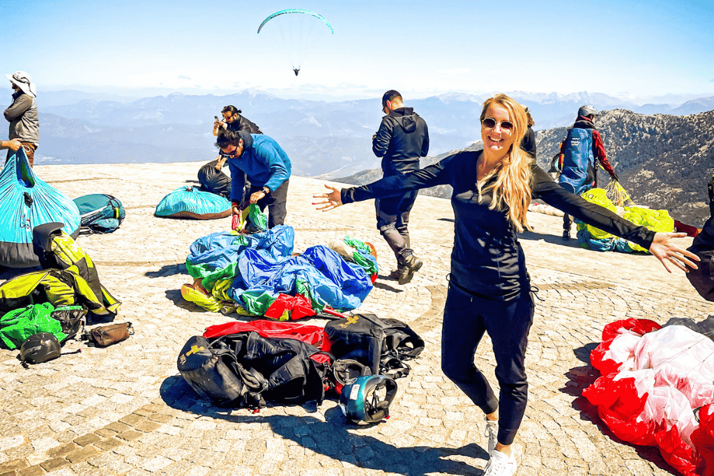 Kate smiles with arms outstretched at the mountaintop launch site in Ölüdeniz, Turkey, surrounded by colorful paragliding gear and a glider soaring above the distant mountain range.