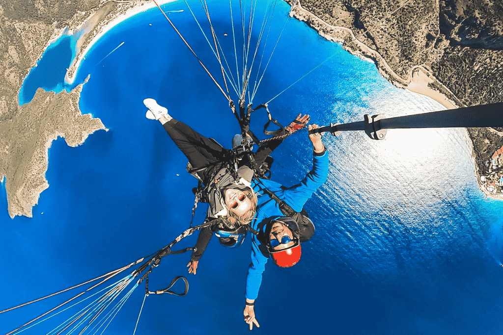 Kate paraglides high above the glittering Blue Lagoon in Ölüdeniz, Turkey, grinning toward the camera with the deep blue sea, curving shoreline, and sunlit water far below.
