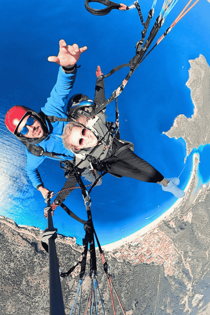 Kate paraglides over the vivid coastline and town of Ölüdeniz, Turkey, smiling as the beach, lagoon, and Mediterranean Sea form a breathtaking backdrop far below.