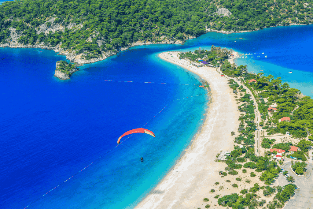 A paraglider sails over the vibrant turquoise waters and golden sands of Ölüdeniz’s famous Blue Lagoon, with lush green hills and anchored boats completing the breathtaking aerial view.