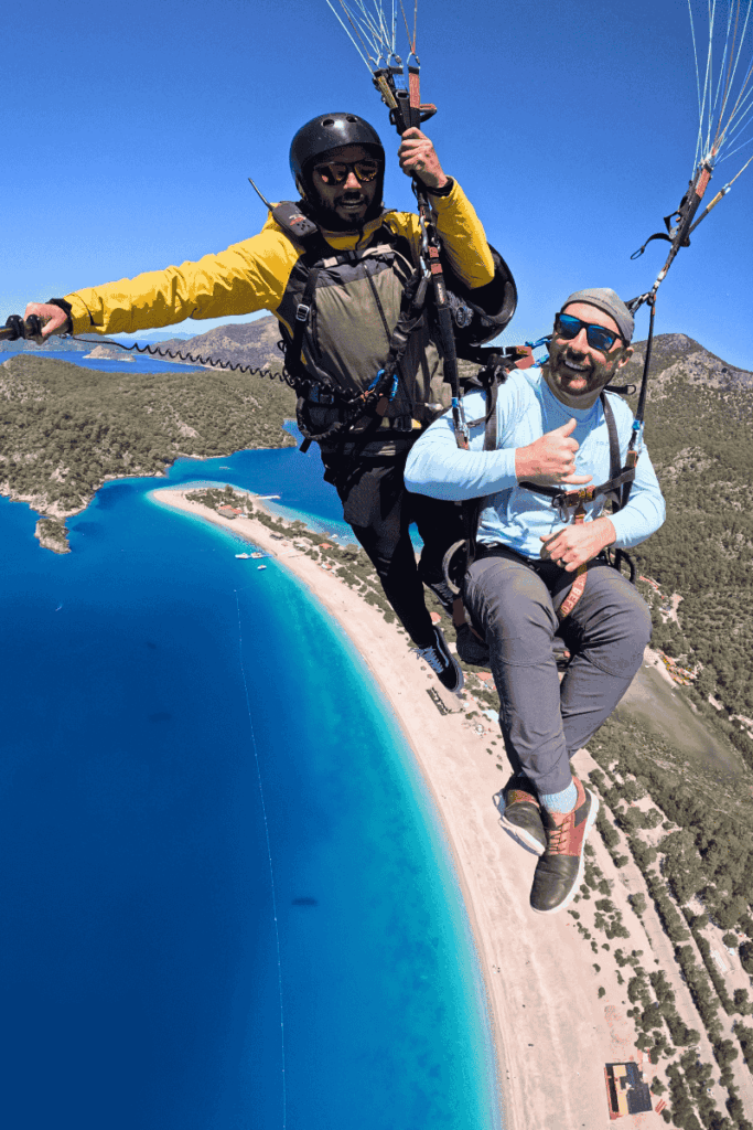Kate’s husband gives a thumbs up while paragliding high above Ölüdeniz’s shoreline, with clear turquoise water and sandy beach far below.