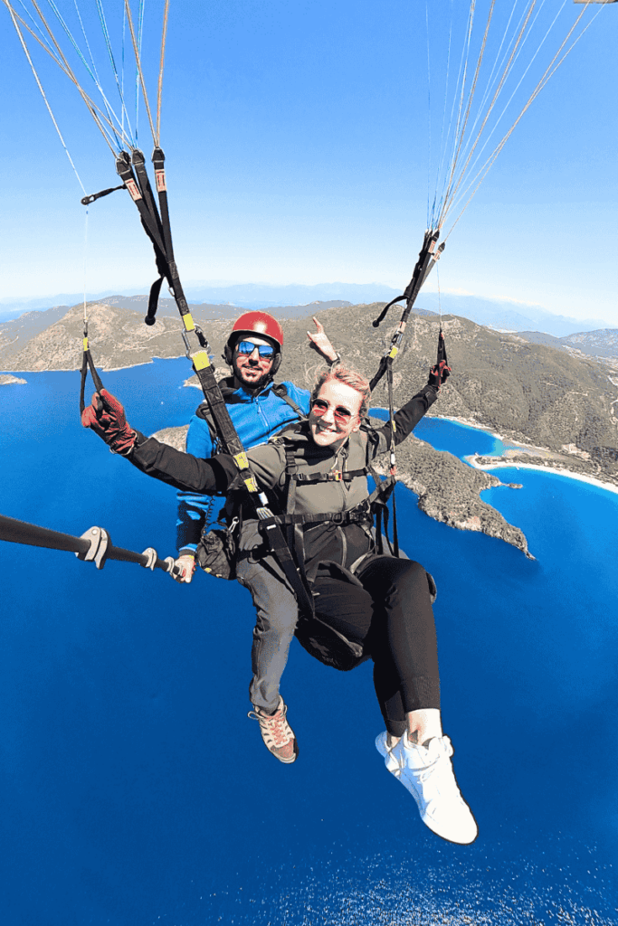 Kate smiles midair during tandem paragliding over the deep blue coast of Ölüdeniz, Turkey, with the Blue Lagoon and green hills stretching out beneath her.