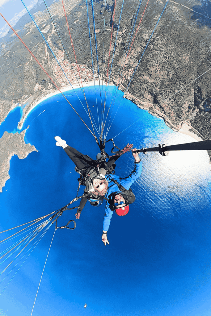 Kate paraglides over the brilliant blue arc of Ölüdeniz bay, smiling as the town, beach, and sparkling sea spread out beneath her and the colorful lines of the canopy fill the sky.