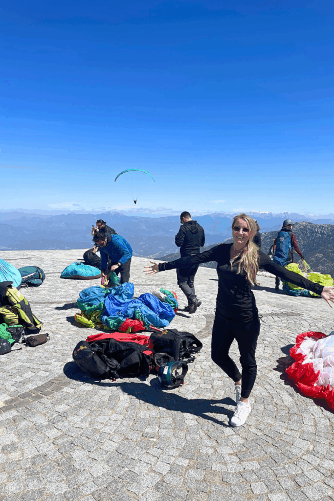 Kate stands smiling with arms open wide on the paragliding launchpad at the summit in Ölüdeniz, Turkey, as instructors prep colorful chutes and a paraglider soars in the background.