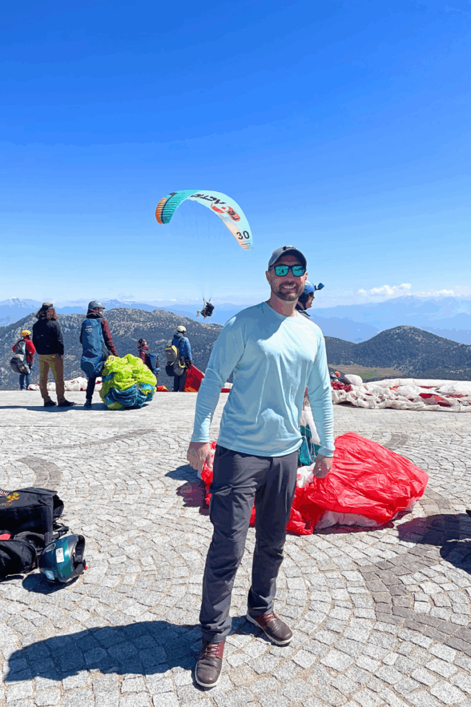 Kate’s husband poses with a smile in front of packed parachutes and fellow paragliders on the mountain top in Ölüdeniz, with one glider already airborne in the clear blue sky behind him.