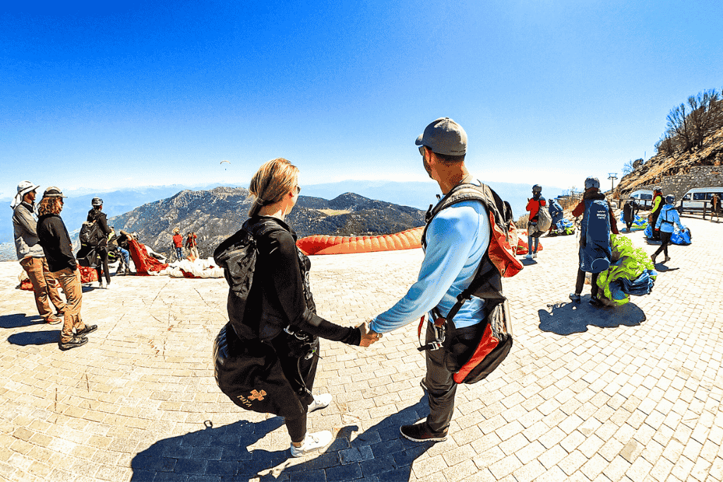 Kate and her husband hold hands while walking toward the launch area on the mountain in Ölüdeniz, surrounded by paragliders preparing their gear under a brilliant blue sky.