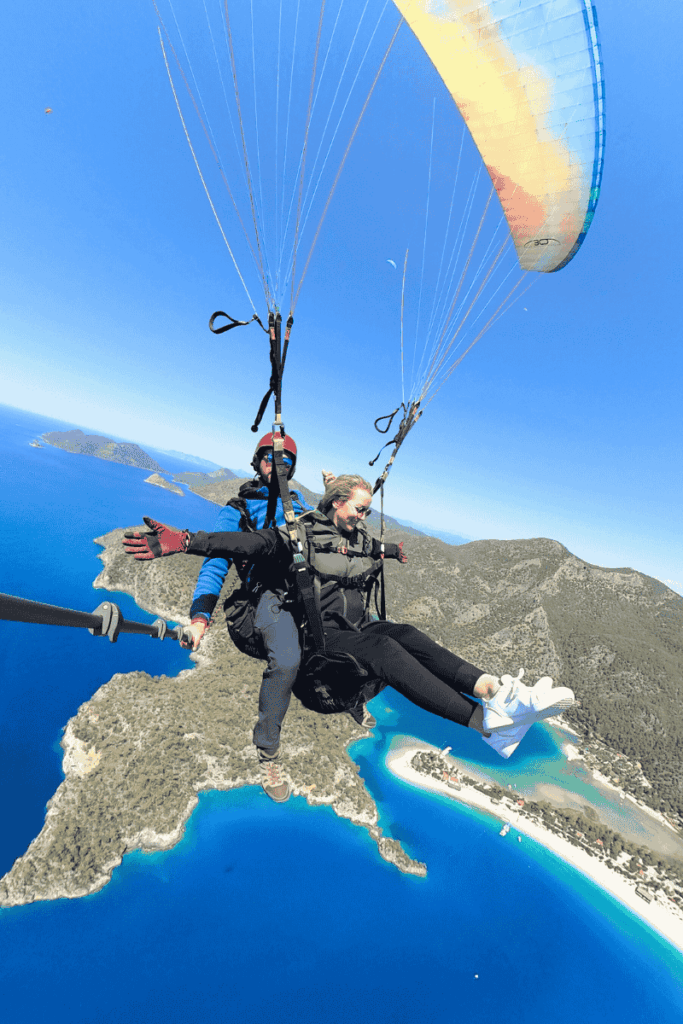 Kate paraglides high above the vivid turquoise coastline and forested cliffs of Ölüdeniz, Turkey, arms outstretched in joy with her pilot as the iconic Blue Lagoon sparkles below.