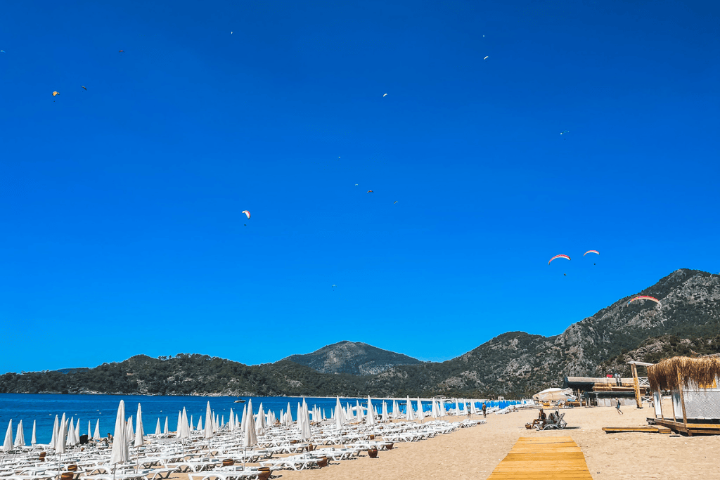 A wide stretch of sandy beach in Ölüdeniz, Turkey, lines up with rows of white sun loungers under a deep blue sky filled with colorful paragliders soaring above the surrounding mountains.