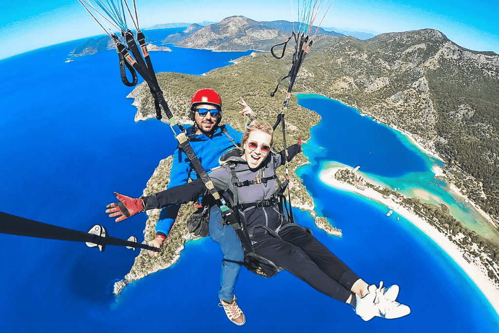 Kate paraglides high above the vibrant turquoise coast of Ölüdeniz, Turkey, smiling with arms outstretched alongside her instructor against a dramatic backdrop of mountains and lagoons.