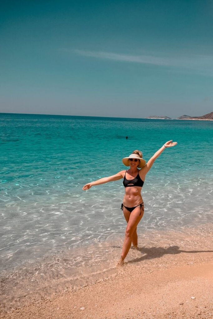 Kate poses joyfully at the edge of the water on a serene beach near Kaş, Turkey, wearing a black bikini and sun hat with sparkling turquoise sea behind her.