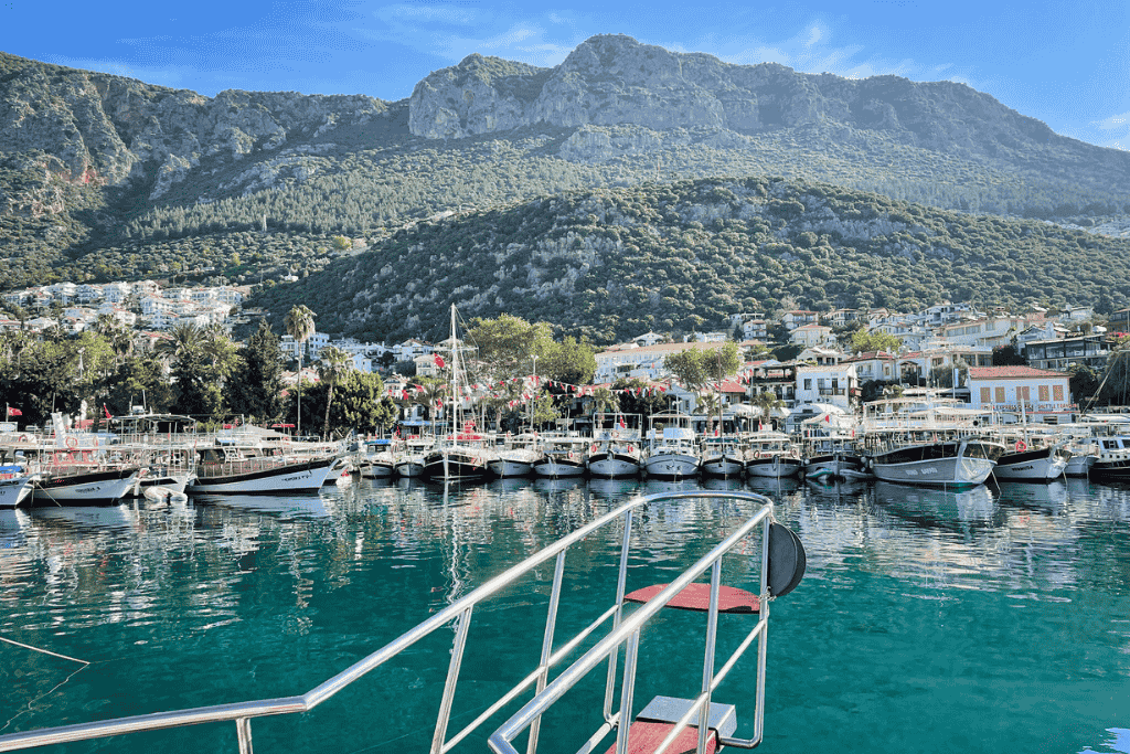 A view from a boat looking toward the charming harbor of Kaş, Turkey, with rows of moored boats, whitewashed buildings, and forested mountains rising in the background under a clear blue sky.