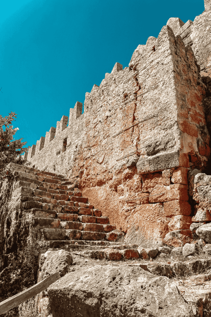 Ancient stone steps lead up the weathered outer wall of Simena Castle, with its crenellated battlements silhouetted against a vibrant blue sky.