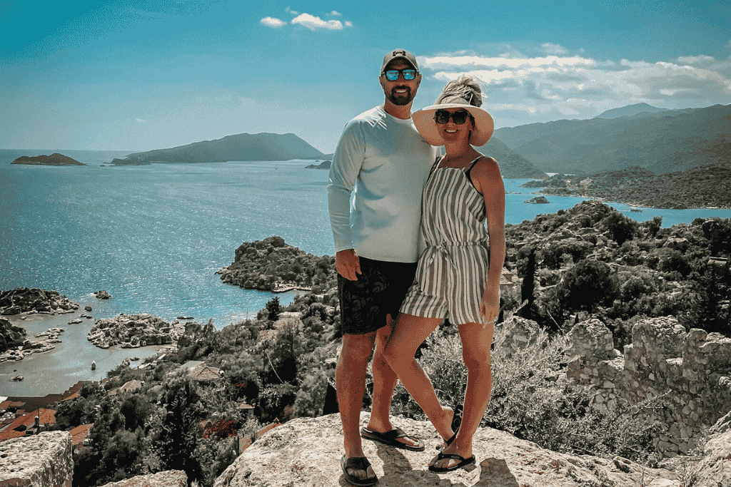 Kate and her husband pose atop Simena Castle with sweeping views of Kaleköy’s rugged coastline, islets, and deep blue Mediterranean waters behind them.