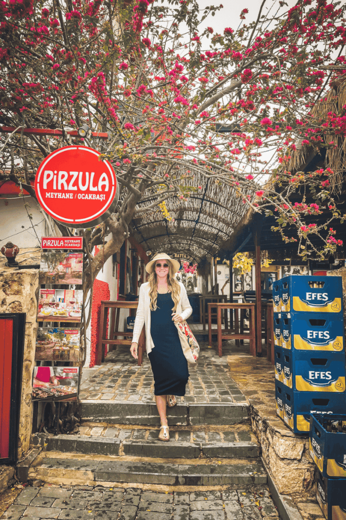 Kate walks past the entrance of Pirzula Meyhane in Kaş, Turkey, under blooming branches and rustic bamboo awnings, with Efes beer crates stacked nearby.