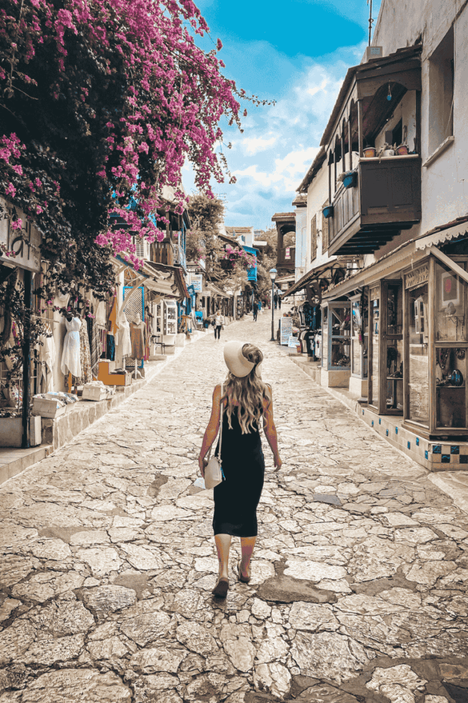 Kate strolls through a quaint stone-paved street in Kaş, lined with whitewashed shops, hanging textiles, and bright bougainvillea under a blue sky.