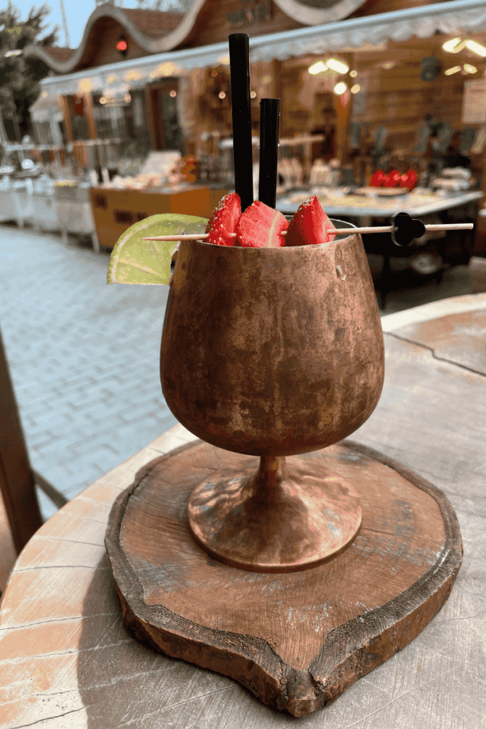 A copper goblet-style cocktail garnished with strawberries and lime sits on a rustic wood slab at an outdoor café in Kaş, Turkey.