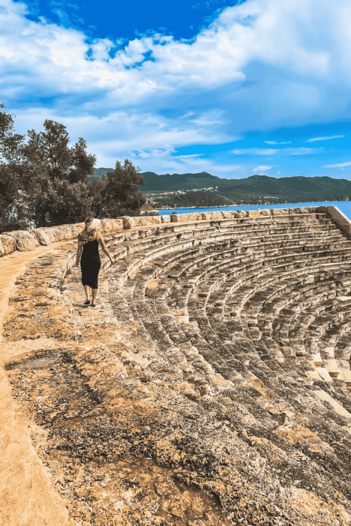 Kate walks along the curved stone seating of the ancient amphitheater at Antiphellos in Kaş, Turkey, with views of the sea and distant mountains.