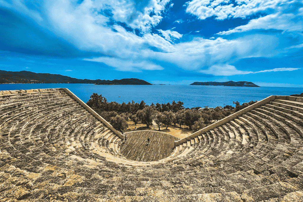 A wide-angle view from the top of the Antiphellos amphitheater in Kaş, Turkey, showcases its semicircular stone seating and a stunning backdrop of the Mediterranean Sea and islands.