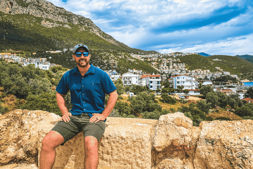 Kate’s husband sits smiling on the stone ledge overlooking the modern town of Kaş, backed by green hills and whitewashed buildings cascading toward the harbor.