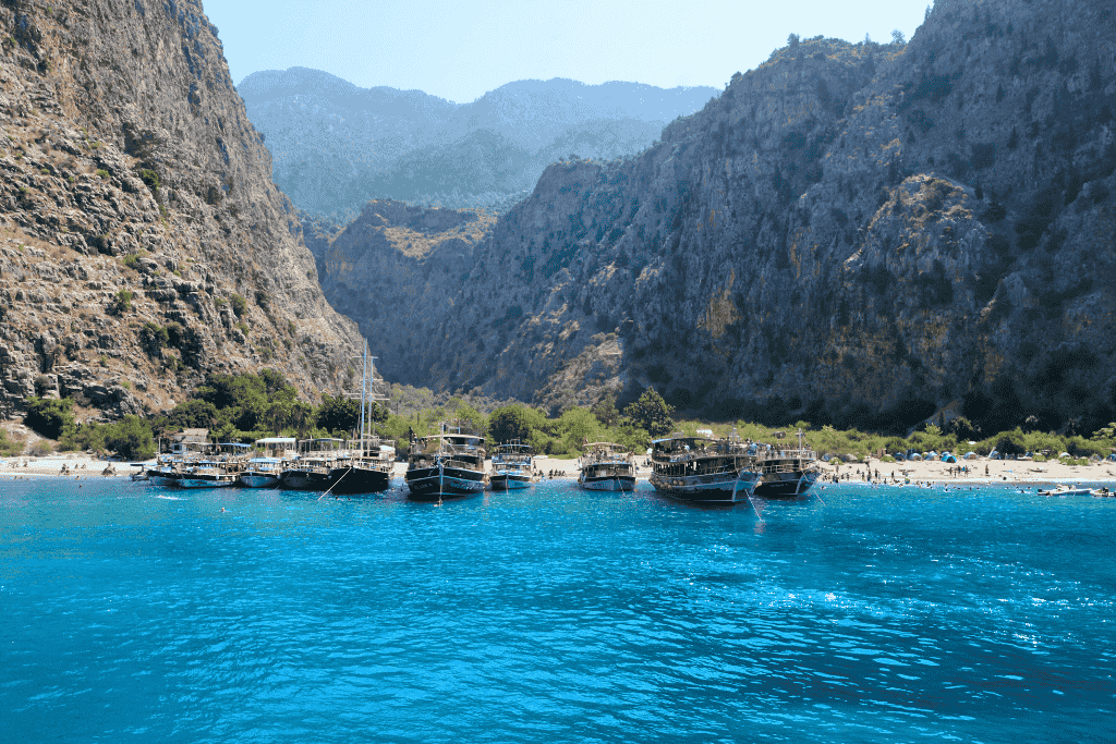 Traditional wooden boats line the shore of Butterfly Valley in Turkey, where a secluded beach meets towering cliffs and vivid turquoise waters under a hazy mountain backdrop.