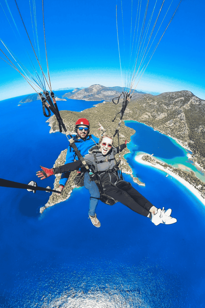 Soaring over the brilliant blue waters of Ölüdeniz, Kate paraglides with a guide, with the lush green peninsula and sandbar curving below them.