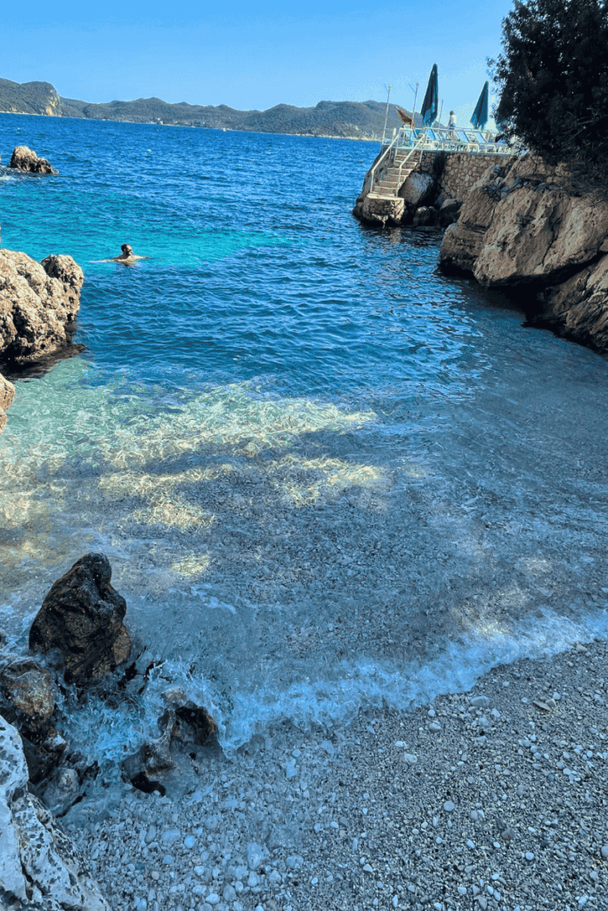 A quiet cove at Small Pebble Beach in Kaş, Turkey, with gentle waves lapping at the pebbled shore and a lone swimmer enjoying the vivid blue sea.