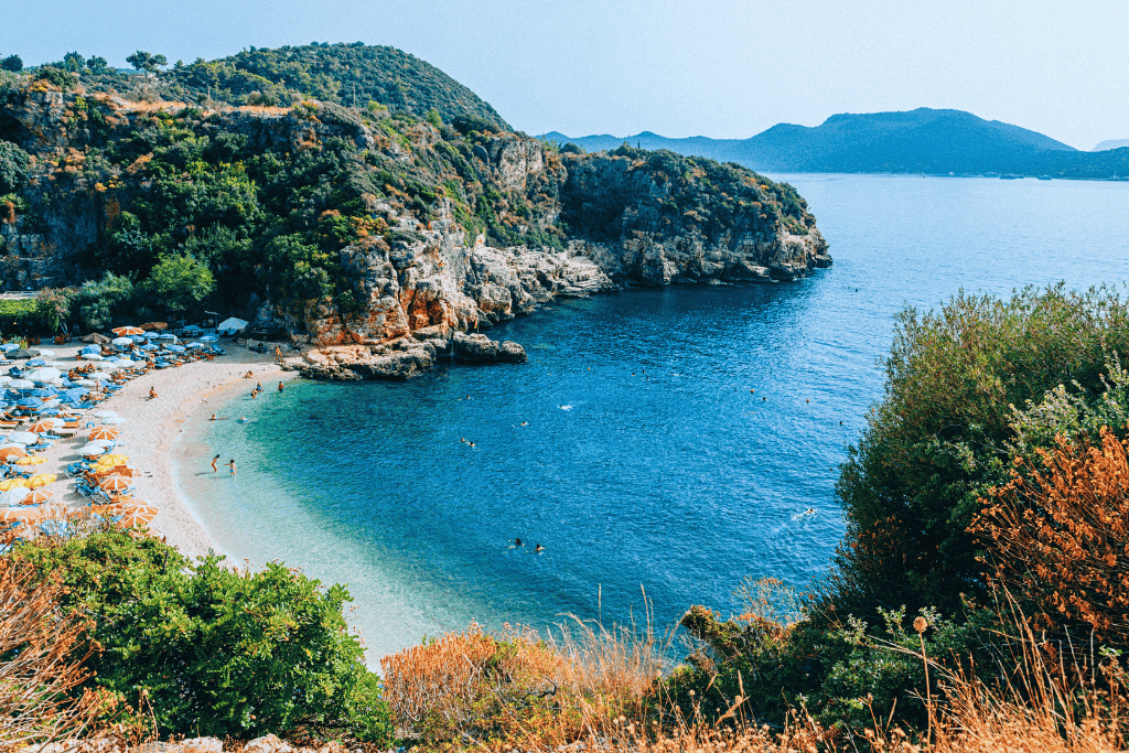 Big Pebble Beach in Kaş, Turkey curves around a scenic bay with colorful umbrellas, swimmers in turquoise water, and rugged cliffs framing the serene Mediterranean coast.