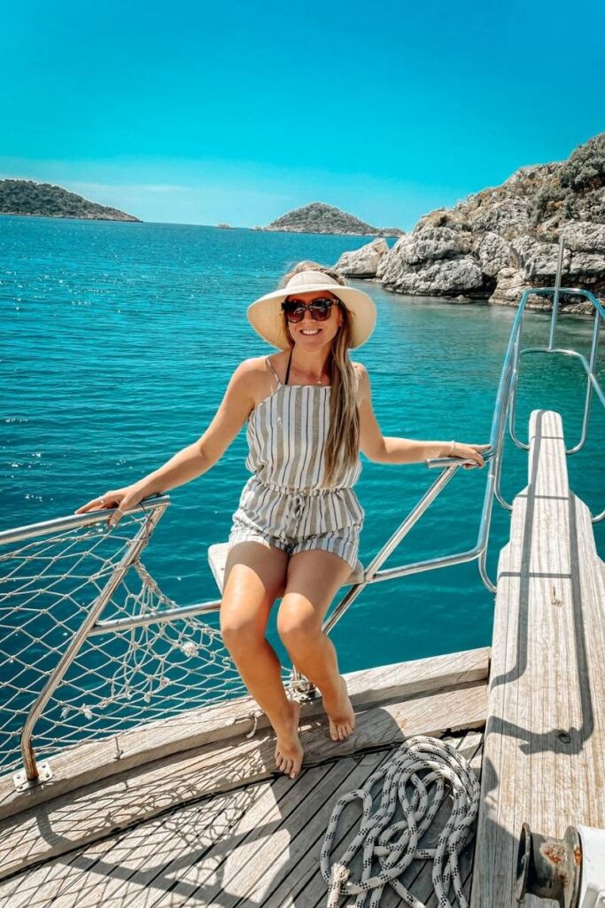 Kate smiles in a striped romper and sunhat while sitting barefoot on the edge of a boat, with turquoise water and rocky islets near Kaş, Turkey in the background.