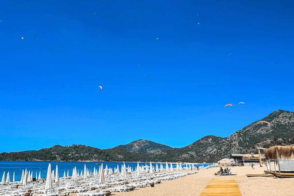 Rows of white loungers and umbrellas line the golden sands of Ouldeniz Beach in Fethiye as colorful paragliders float through the clear blue sky above the surrounding mountains.
