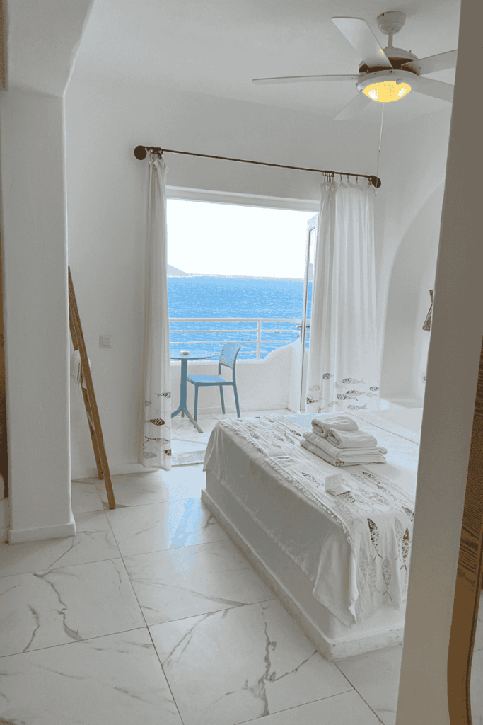 Bright, minimalist hotel room with white decor and an open balcony door offering a clear view of the Aegean Sea, a small table and chair set visible outside.
