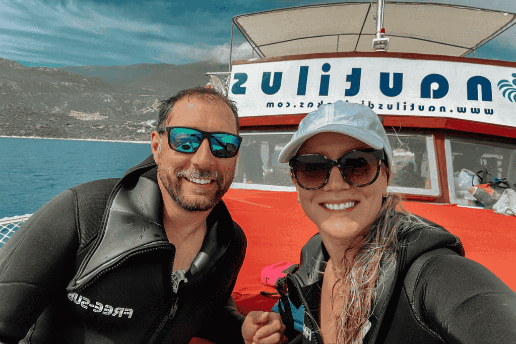 Kate and her husband smile in wetsuits for a post-dive selfie in front of the red and white Nautilus dive boat, with Kaş’s rocky coastline behind them.