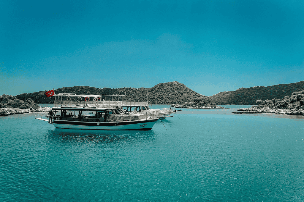 A traditional Turkish gulet boat with a red Turkish flag floats in calm turquoise waters, surrounded by the lush, hilly islets near Kaş, Turkey under a clear blue sky.