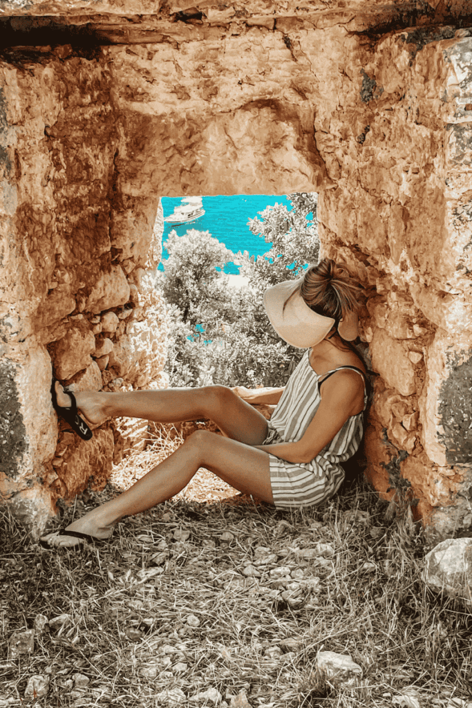Kate relaxes in a stone window nook of Simena Castle, looking out over the turquoise sea and anchored boats near Kaleköy, Turkey.