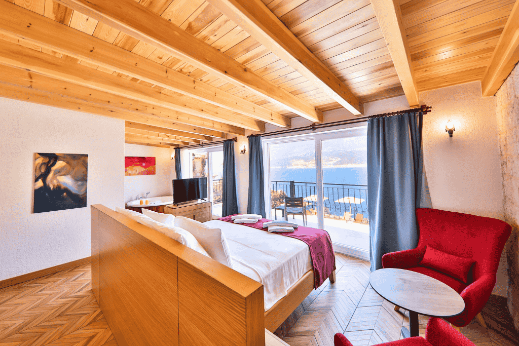 Warm-toned hotel suite in Kaş with wood ceilings, sea-view balcony, and cozy red armchairs by the window.