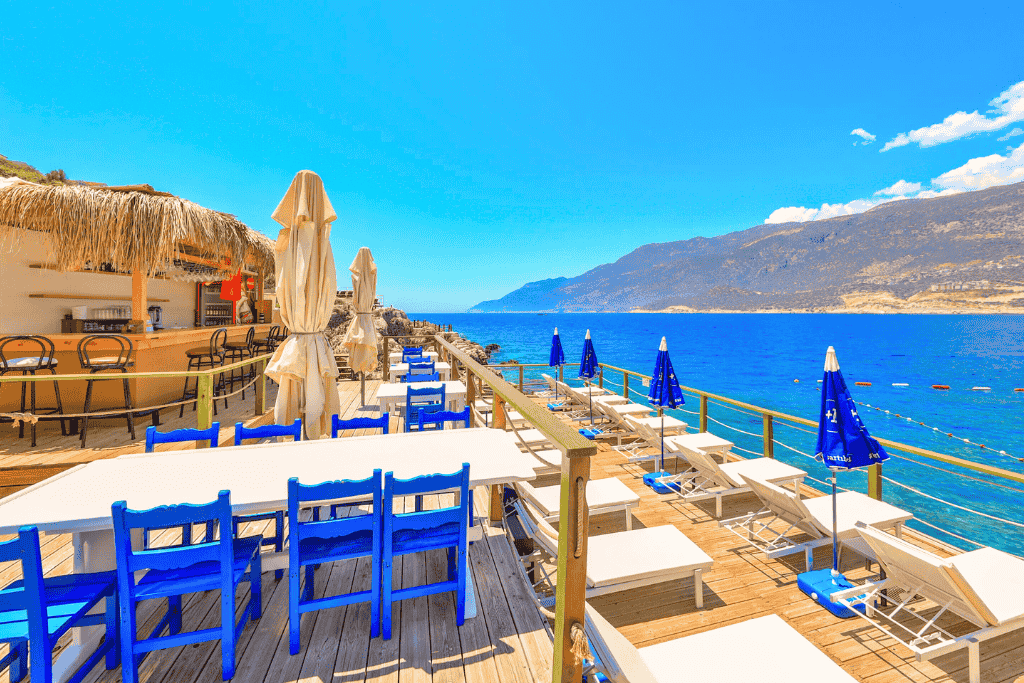 Beachfront deck at a Kaş hotel with blue chairs, sun loungers, and umbrellas overlooking the bright turquoise sea and mountains.