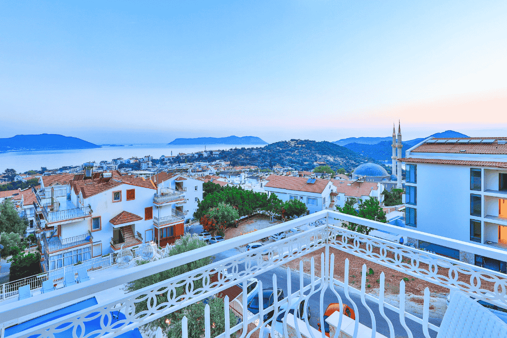 Peaceful hotel balcony view in Kaş at dusk, overlooking whitewashed buildings, a mosque, and distant islands in the sea.