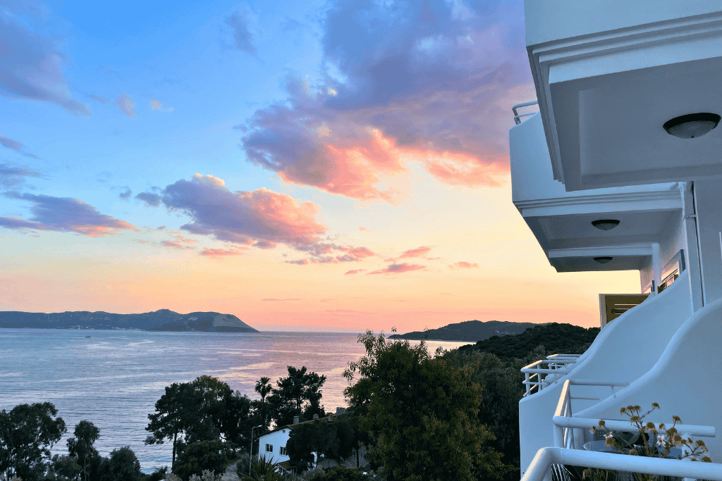 View of a colorful sunset over the sea from a hotel balcony in Kaş, with nearby islands and lush greenery below.