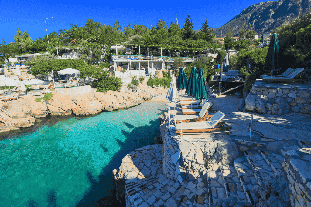 Rocky seaside terrace in Kaş with sun loungers and umbrellas overlooking a crystal-clear turquoise cove surrounded by lush greenery.