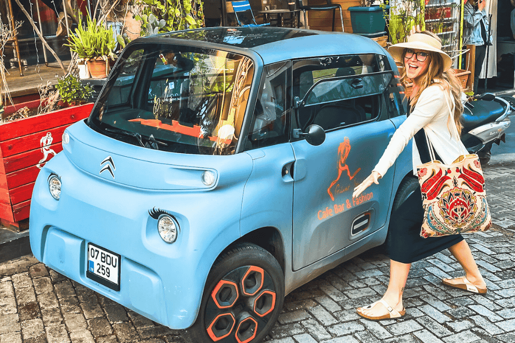Smiling, Kate in Kaş points at a small blue Citroën Ami with orange wheels and cafe branding, parked on a cobblestone street.