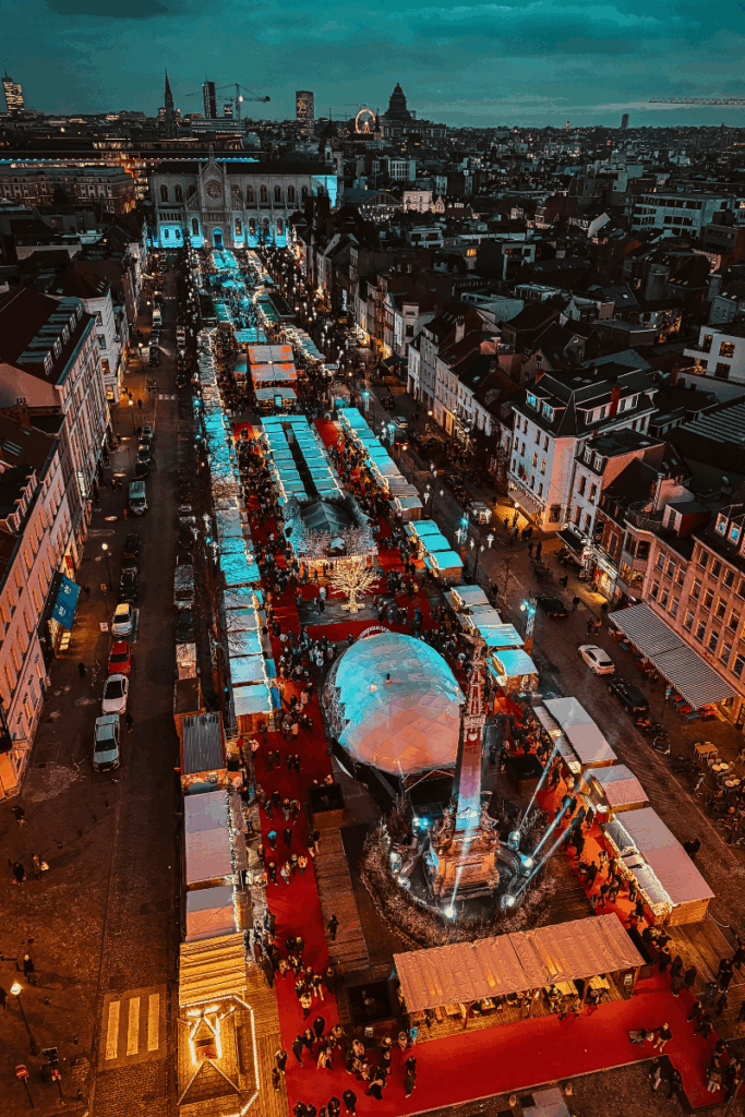 Vibrant aerial view of Brussels Christmas Market at night, with glowing market stalls, red carpets, a central monument, and festive lights stretching toward the illuminated church and city skyline.
