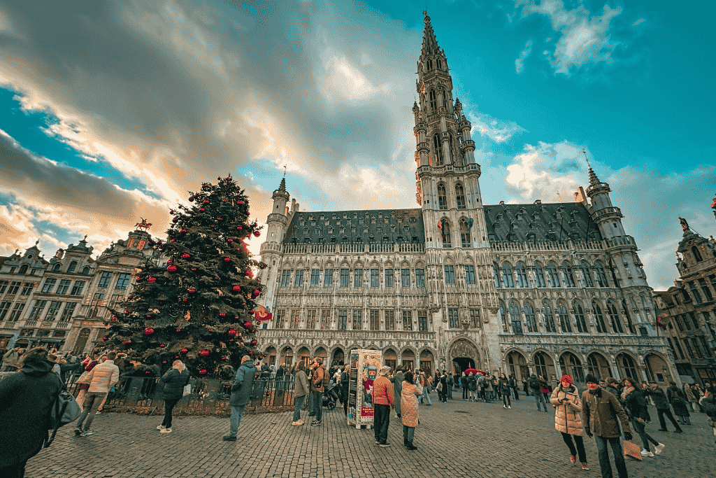 A large, decorated Christmas tree stands in the bustling Grand Place square, framed by the ornate Gothic architecture of the Town Hall and surrounded by tourists enjoying the festive season.