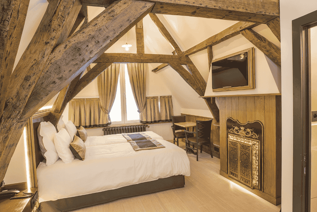 Cozy, rustic-chic hotel room at Hotel Agora Brussels Grand Place featuring exposed wooden beams, a plush bed, ornate fireplace, and warm neutral tones.

