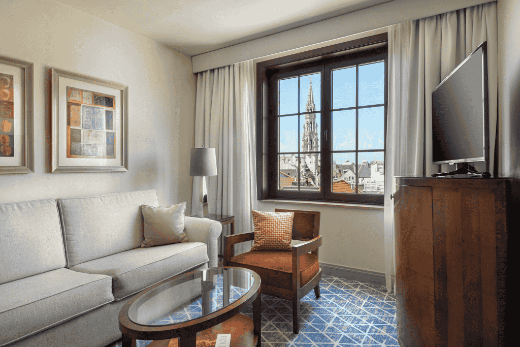 Elegant hotel room at Warwick Brussels Grand Place with a cozy sitting area, neutral-toned decor, and a stunning view of the Town Hall spire through the window.

