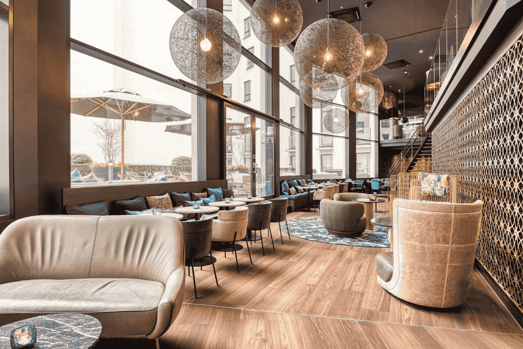 Chic and modern lounge area at Motel One Brussels, featuring high ceilings, floor-to-ceiling windows, stylish globe pendant lights, and cozy seating in neutral and blue tones.