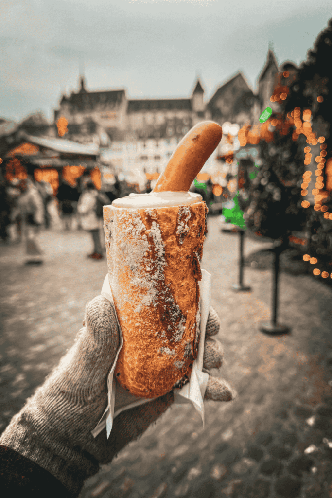 A gloved hand holds a chimney roll filled with creamy sauce and topped with a sausage at a European Christmas market with festive lights in the background.
